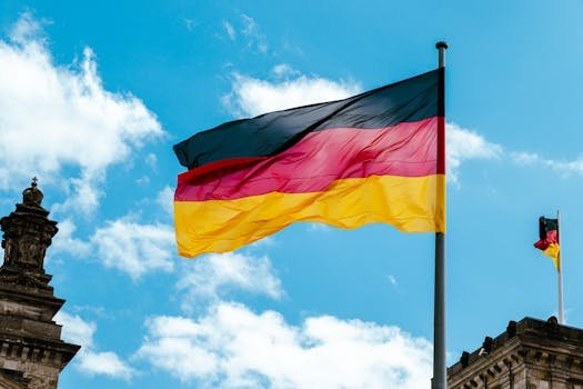Vibrant German flag waving on a flagpole against a bright blue sky with clouds.
